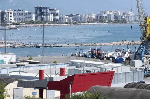 People gather on the beach as a migrant center is seen in the foreground in the port of Shengjin, northwest Albania, Thursday, July 25, 2024. Migrants rescued at sea while attempting to reach Italy are likely to see themselves transported to Albania from next month while their asylum claims are processed, under a controversial deal in which the small Balkan country will host thousands of asylum-seekers on Italy's behalf. (AP Photo/Vlasov Sulaj)