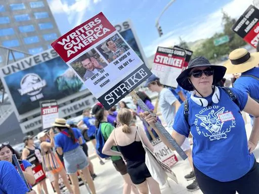 Writer Julie Benson holds a picket sign with an image of the late actor and comic Paul Reubens, dressed as his character Pee-wee Herman, outside Universal Studios on Tuesday, Aug. 1, 2023, in Universal City, Calif. The actors strike comes more than two months after screenwriters began striking in their bid to get better pay and working conditions. (AP Photo/Rick Taber)